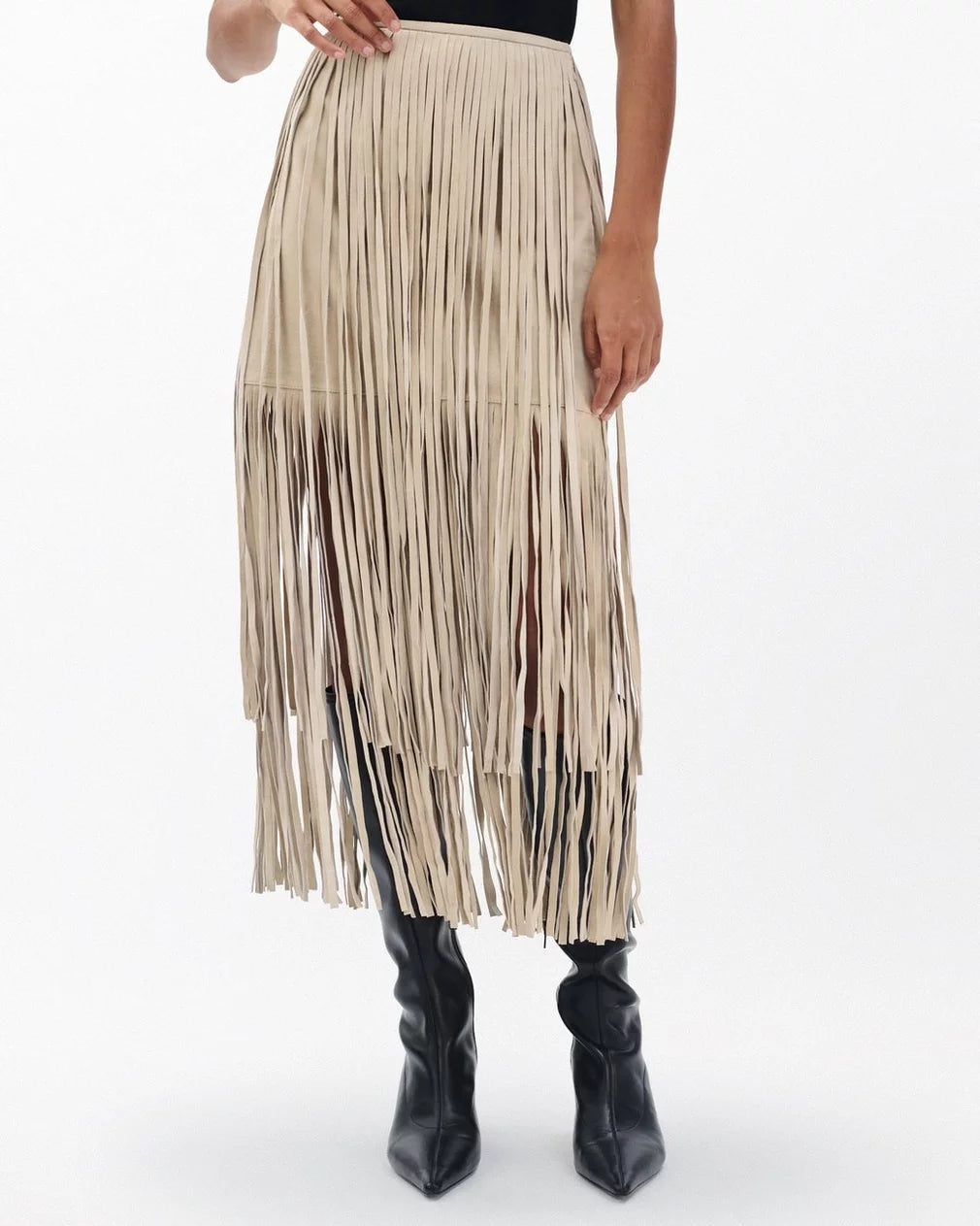 A person wears the Rag & Bone jasmine fringe skirt—a beige, high-waisted suede skirt with long fringe—paired with black knee-high boots, standing against a plain white background.