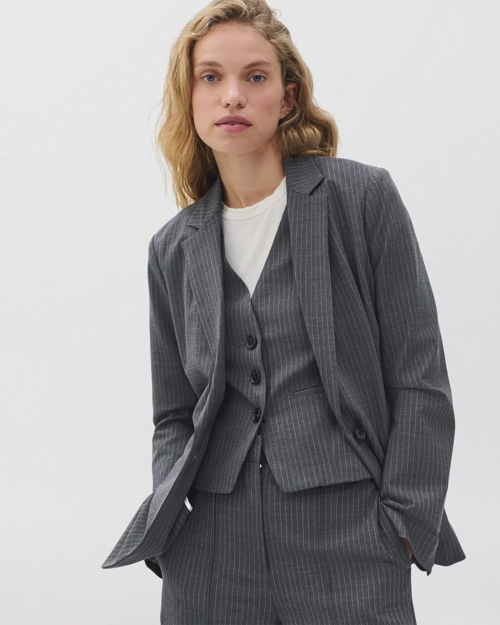 A woman with wavy blonde hair wears the Rag & Bone jennifer striped combo blazer over a white top as a sharp suit, standing confidently against a light background.