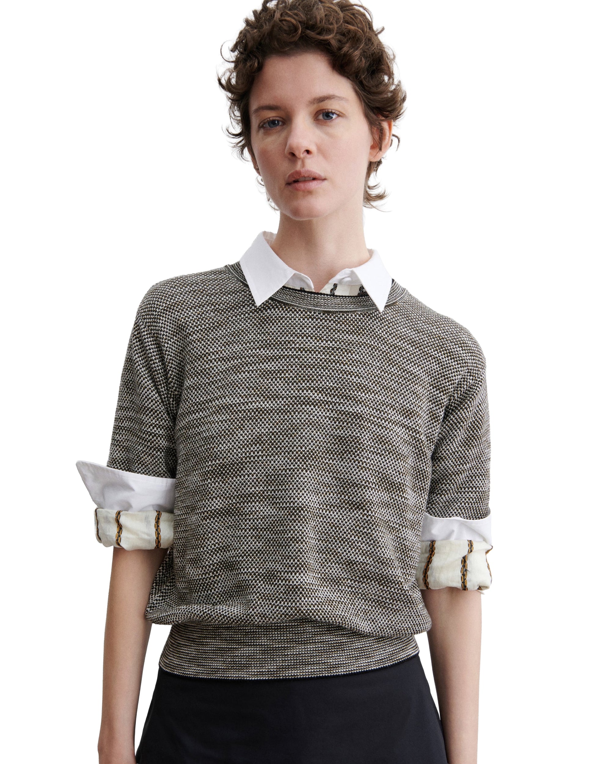 A person with short, curly brown hair wears the Rachel Comey Jeram Top—a textured cotton tweed knit crewneck—over a white collared shirt with rolled-up sleeves and a black skirt, standing against a white background.