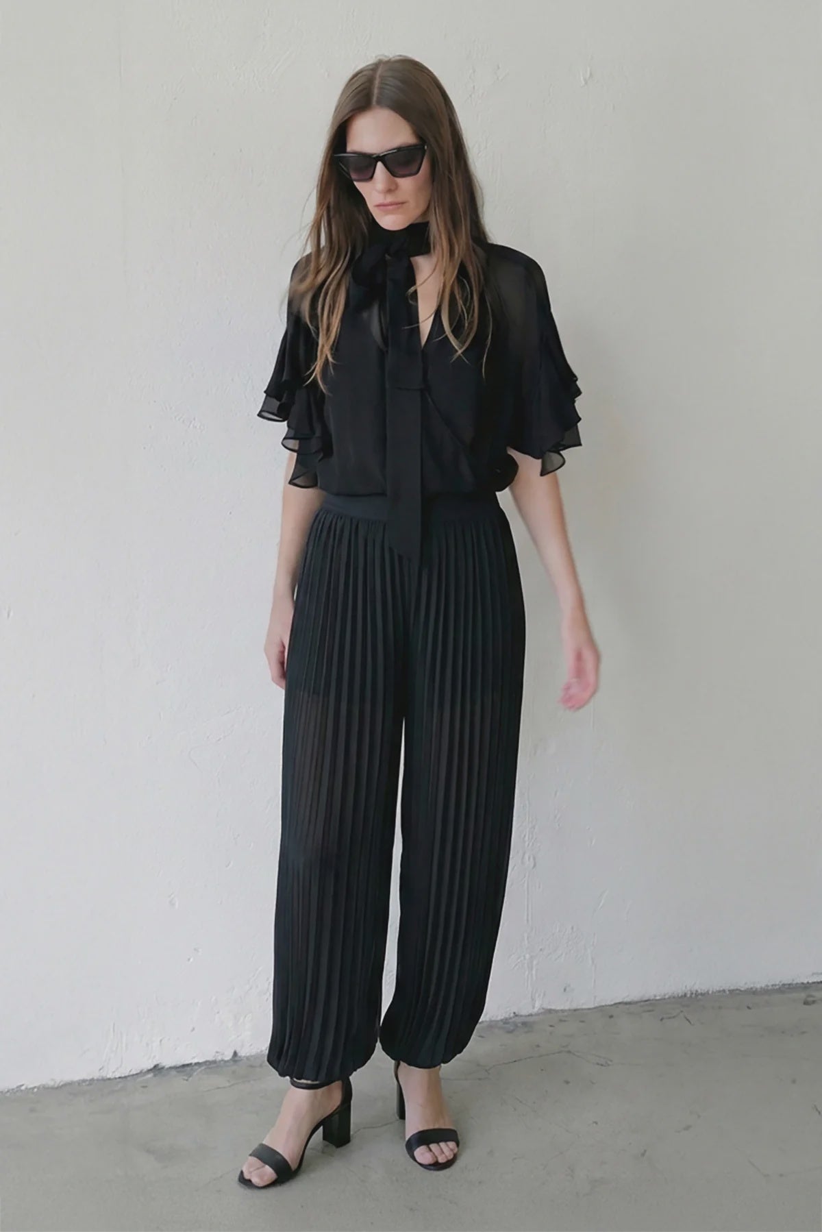 A woman wearing black sunglasses, a sheer black blouse with bow tie neck and flutter sleeves, Le Superbe Pleated Harem Pants, and black heeled sandals stands against a plain white wall.