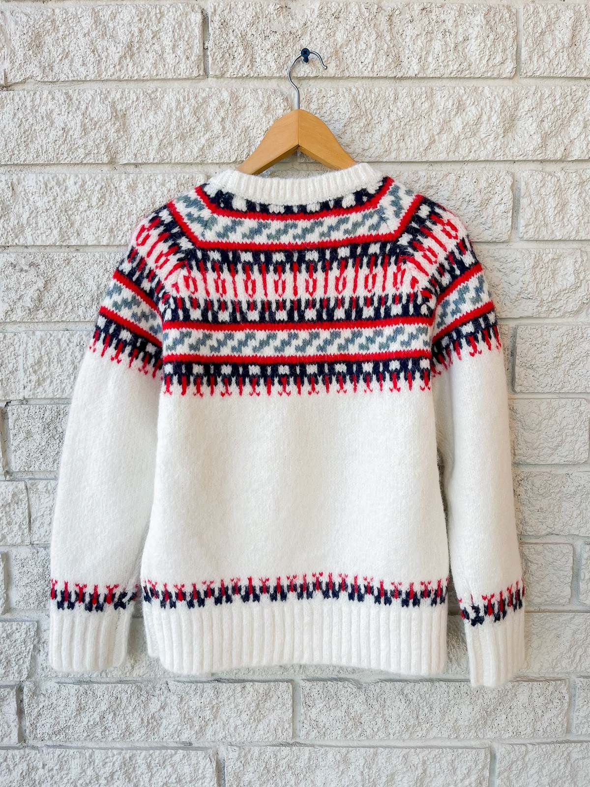 The Josie Sweater by Hunter Bell NYC—a white knit made from wool blend and recycled polyester with red and navy geometric patterns—hangs on a wooden hanger against a white brick wall.