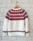 The Josie Sweater by Hunter Bell NYC—a white knit made from wool blend and recycled polyester with red and navy geometric patterns—hangs on a wooden hanger against a white brick wall.