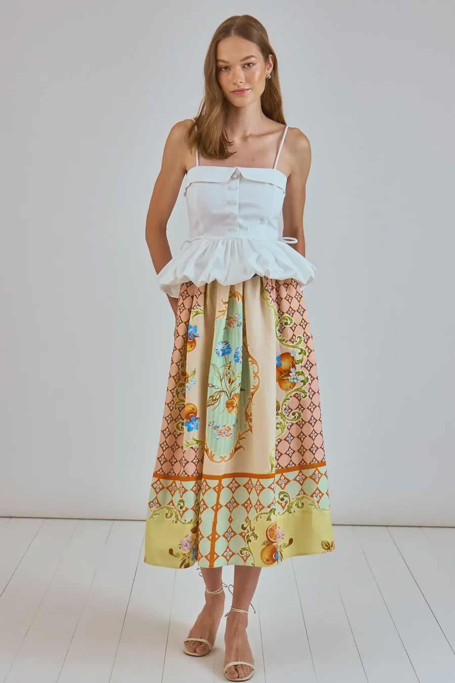 In a minimalistic room, a woman wears Hunter Bell NYC's Tatum Skirt in vibrant Gasparilla Gardens print with a white sleeveless peplum top. Her light brown hair frames her neutral expression, and she pairs the ensemble with strappy sandals, ideal for any spring wardrobe.