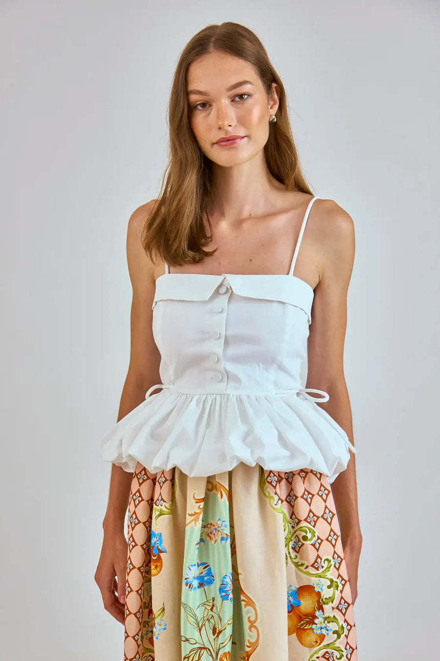 A woman with long brown hair wears a white, strapless top featuring buttons and a ruffled hem. Her Hunter Bell NYC Tatum Skirt displays the vibrant Gasparilla Gardens print, showcasing colorful floral and geometric patterns as she stands against a plain background.