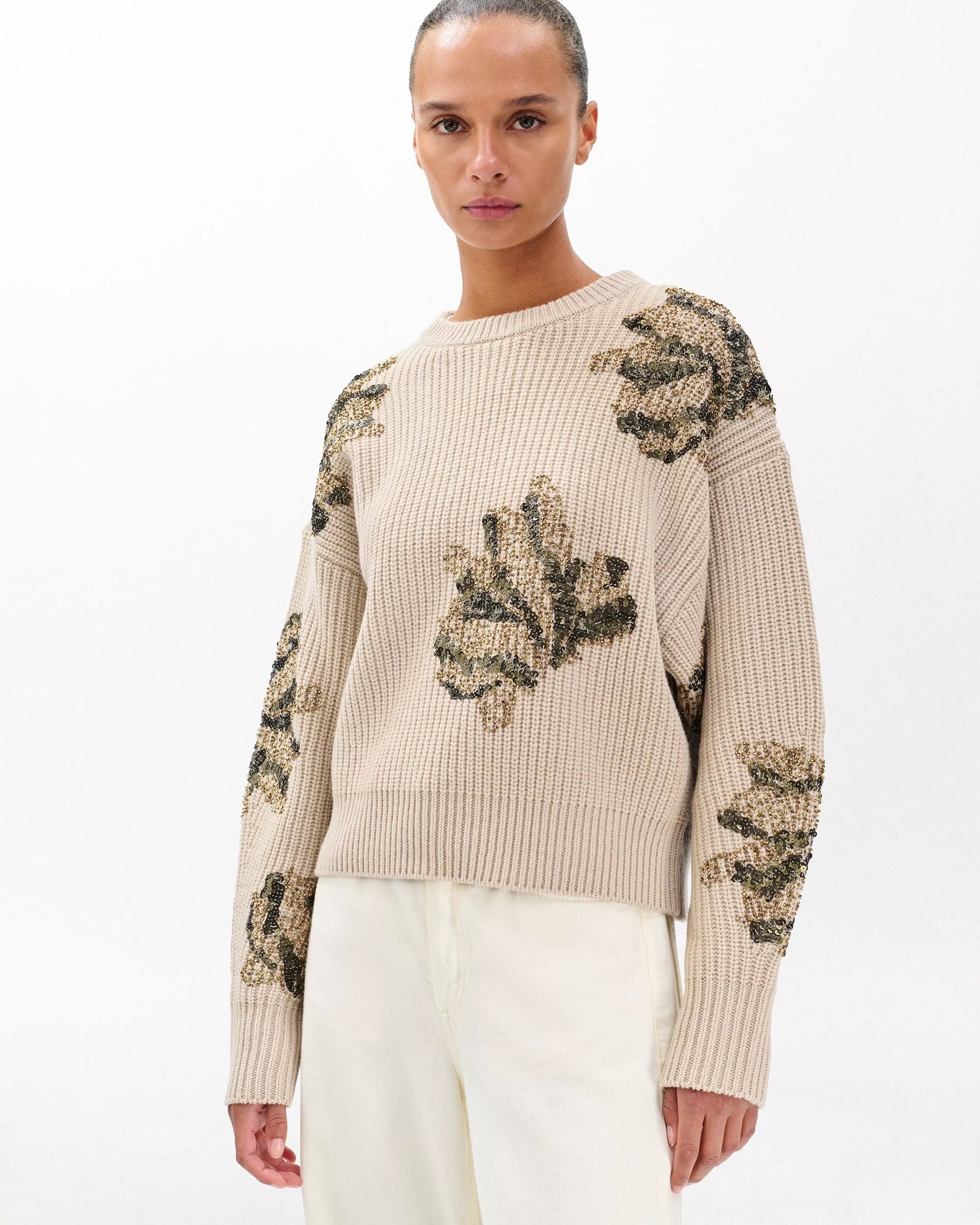 A woman wears the Rag & Bone julie sequin wool sweater in beige with gold, black, and green floral embroidery, paired with off-white pants, standing against a plain white background.
