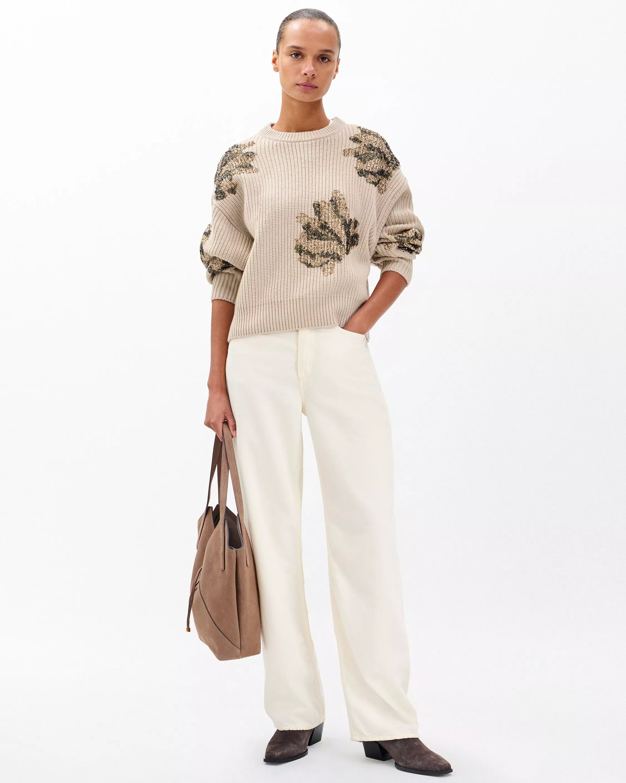 A woman stands against a white background in the Rag & Bone julie sequin wool sweater, featuring floral patterns and green sequins, paired with white wide-leg pants, brown boots, a taupe handbag, and sleekly pulled-back hair.