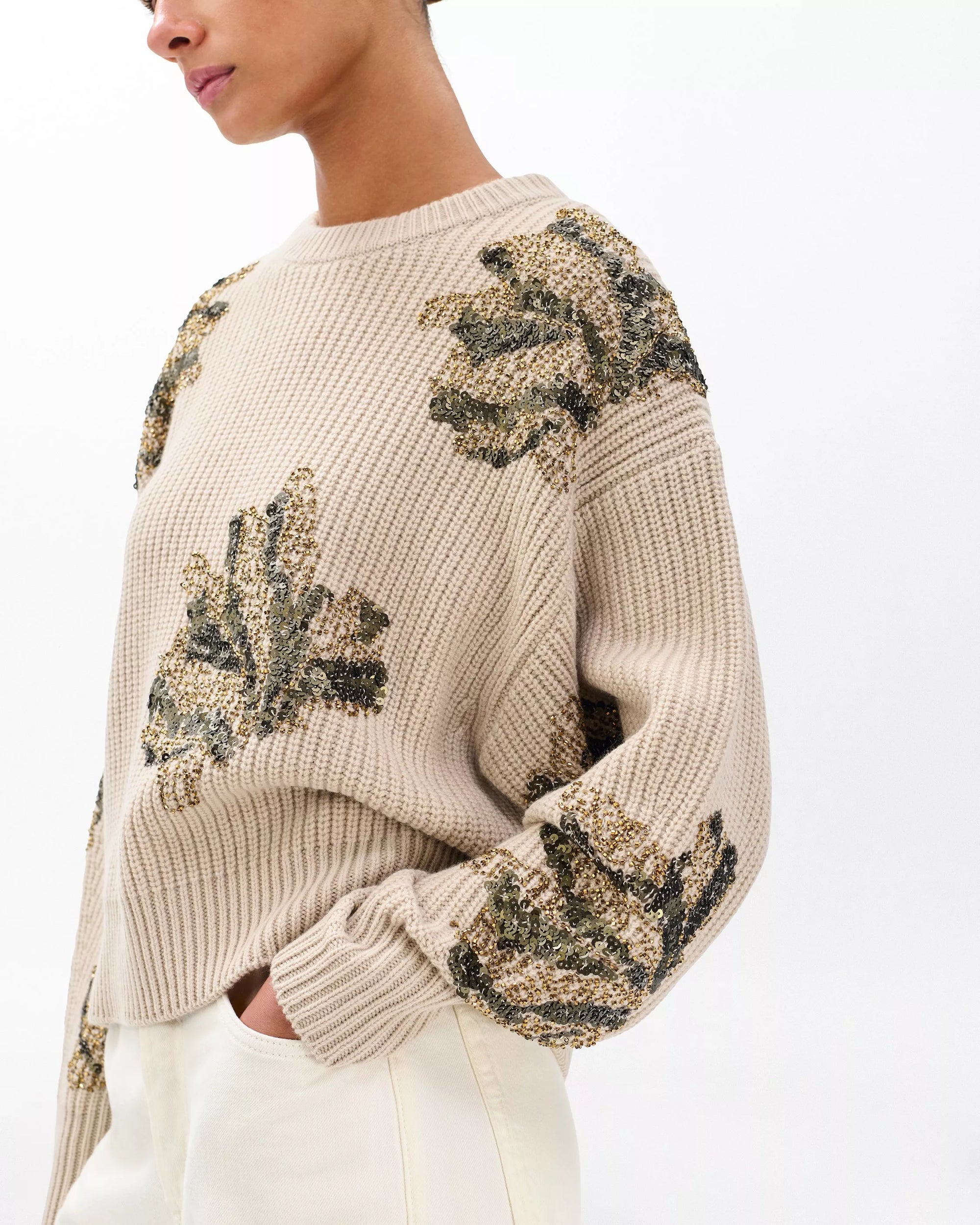 A person wearing the Rag & Bone julie sequin wool sweater—an oversized beige knit with green and gold floral embroidery—paired with light pants, stands against a plain white background.