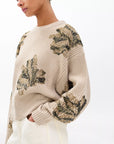 A person wearing the Rag & Bone julie sequin wool sweater—an oversized beige knit with green and gold floral embroidery—paired with light pants, stands against a plain white background.