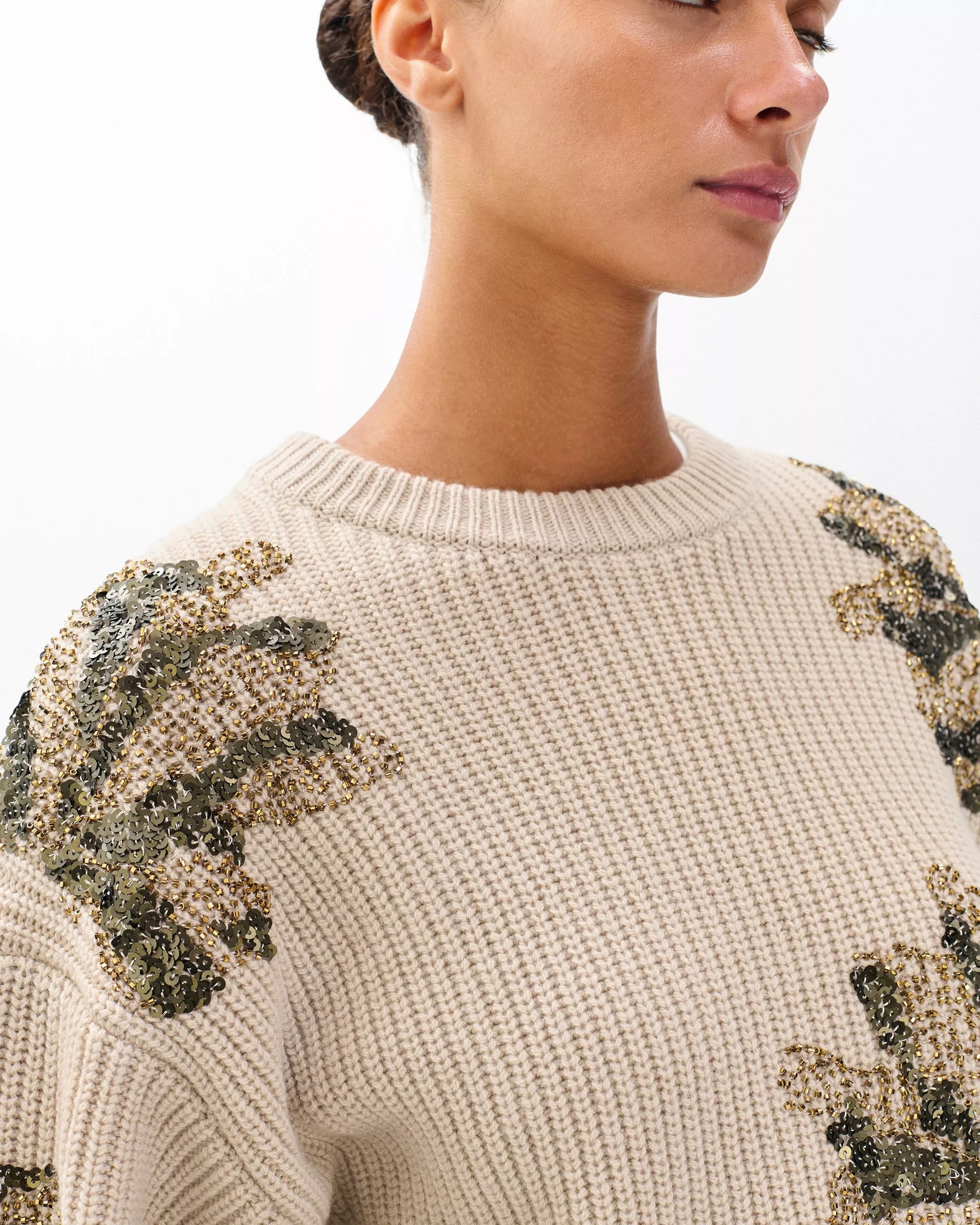 A woman stands against a white background wearing the Rag & Bone julie sequin wool sweater—a beige, chunky knit crewneck featuring green and gold sequin floral embellishments on the shoulders and sleeves.
