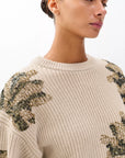 A woman stands against a white background wearing the Rag & Bone julie sequin wool sweater—a beige, chunky knit crewneck featuring green and gold sequin floral embellishments on the shoulders and sleeves.