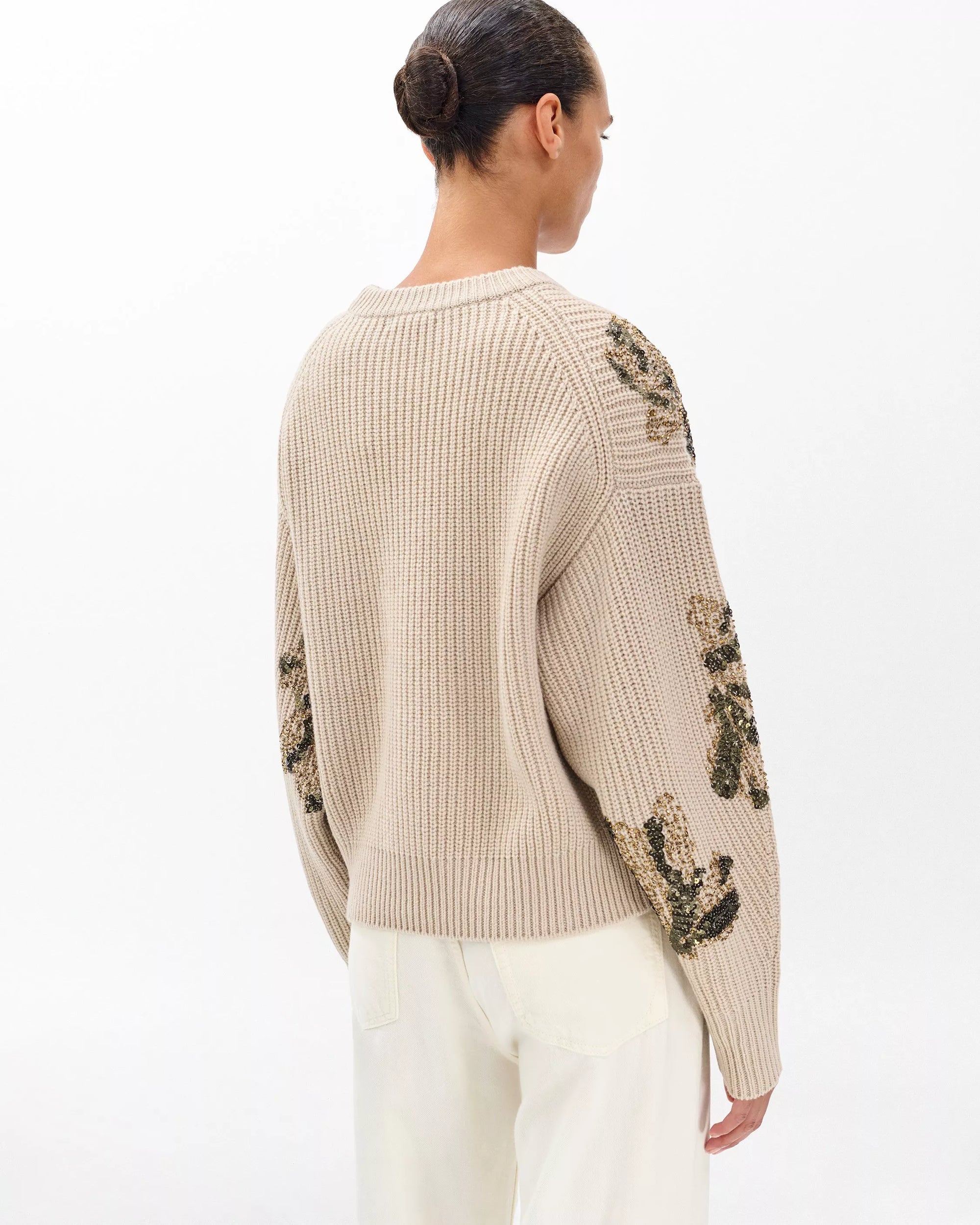 A woman with dark hair in a bun models the Rag & Bone julie sequin wool sweater, featuring floral-embroidered sleeves, paired with light pants. She stands with her back to the camera against a plain white background.