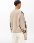A woman with dark hair in a bun models the Rag & Bone julie sequin wool sweater, featuring floral-embroidered sleeves, paired with light pants. She stands with her back to the camera against a plain white background.