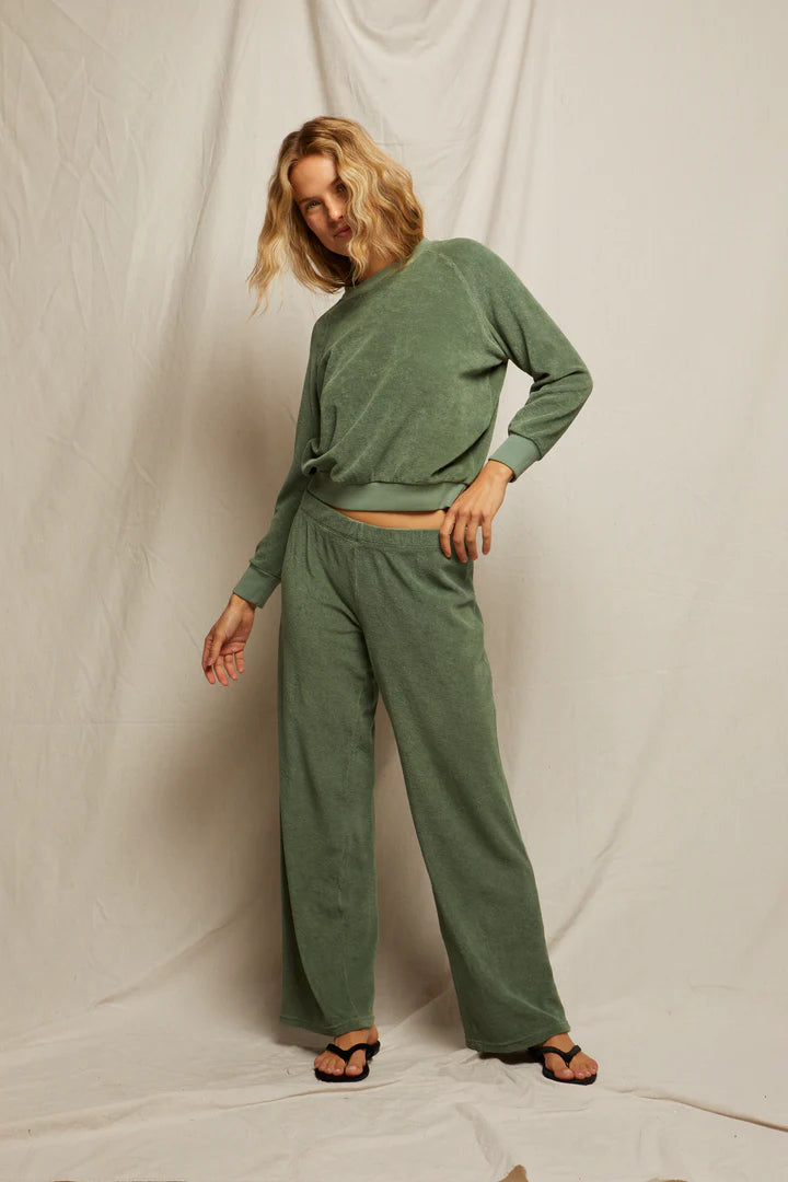 A person with shoulder-length blonde hair wears the Perfectwhitetee Saylor Loop Terry Sweatshirt in sage green with matching wide-leg pants and black sandals, standing against a plain fabric backdrop.