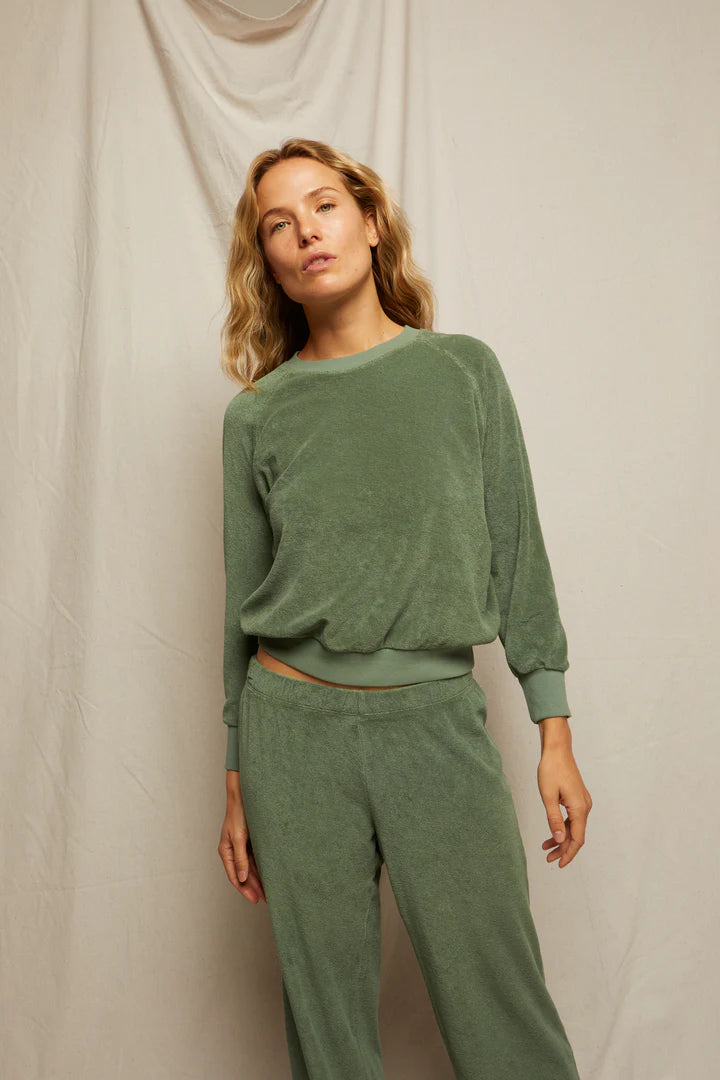 A woman with wavy blonde hair stands against a neutral fabric backdrop, wearing the Perfectwhitetee Saylor Loop Terry Sweatshirt in matching sage green with long sleeves and soft loop terry fabric pants.
