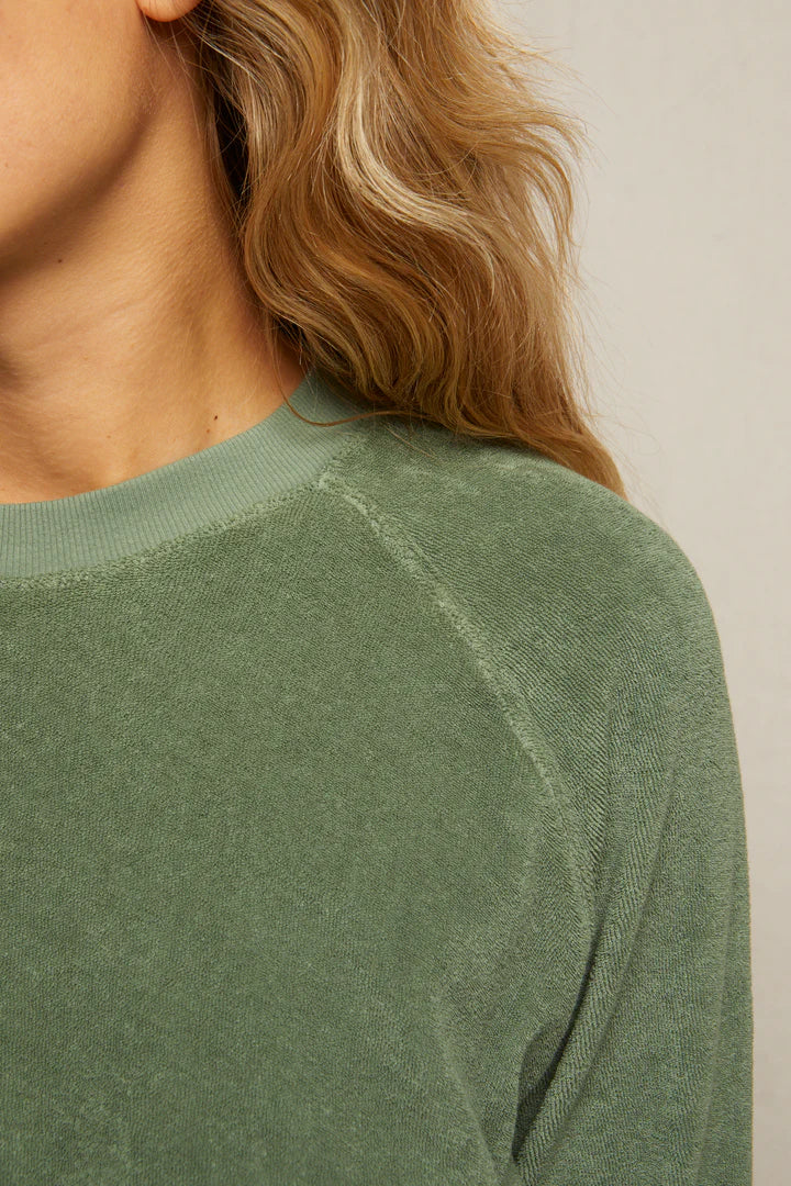 Close-up of a person with long, wavy blonde hair wearing the Perfectwhitetee Saylor Loop Terry Sweatshirt in sage green, showing only the side of the face, neck, shoulder, and upper chest against a plain background.