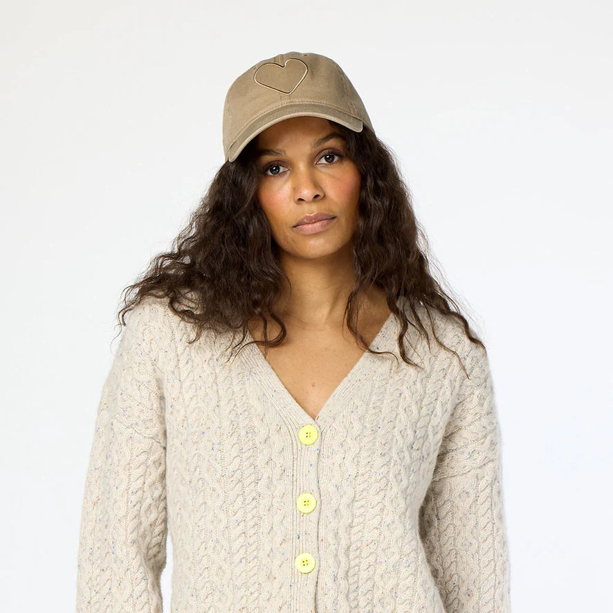 A person with long curly hair wearing a beige knit sweater with yellow buttons and a beige Kerri Rosenthal Imperfect Heart Hat stands against a plain background, exuding a neutral expression.