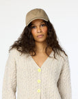 A person with long curly hair wearing a beige knit sweater with yellow buttons and a beige Kerri Rosenthal Imperfect Heart Hat stands against a plain background, exuding a neutral expression.