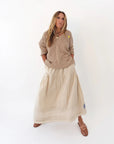 A woman with long hair stands against a white background wearing the Kerri Rosenthal Summerland V Neck Sweater, paired with a long cream skirt and brown sandals. Her hands are in her pockets.