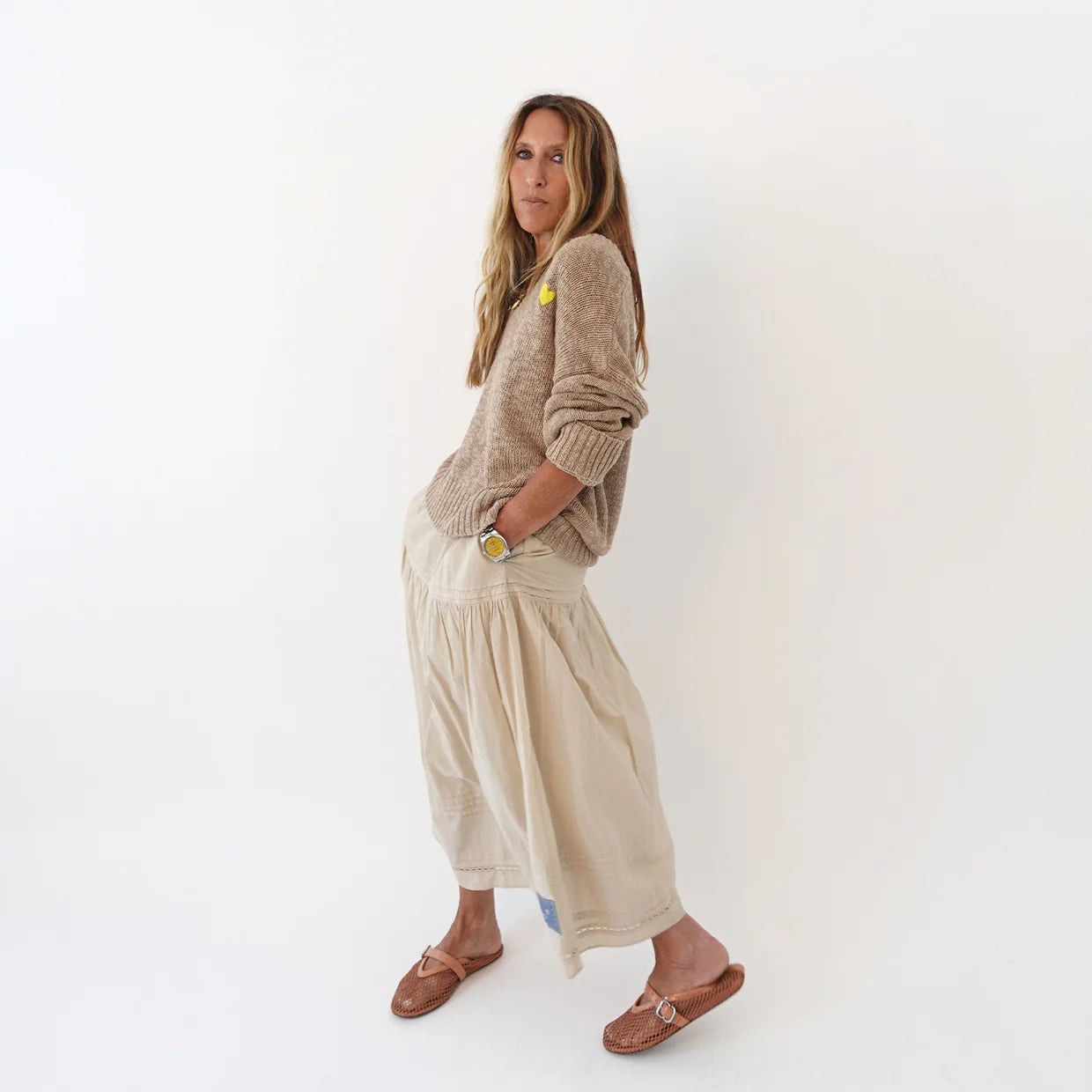 A woman with long brown hair stands sideways against a white background, wearing the Kerri Rosenthal Summerland V Neck Sweater, a beige ankle-length skirt, brown sandals, and a yellow watch. Her hands are in her skirt pockets.