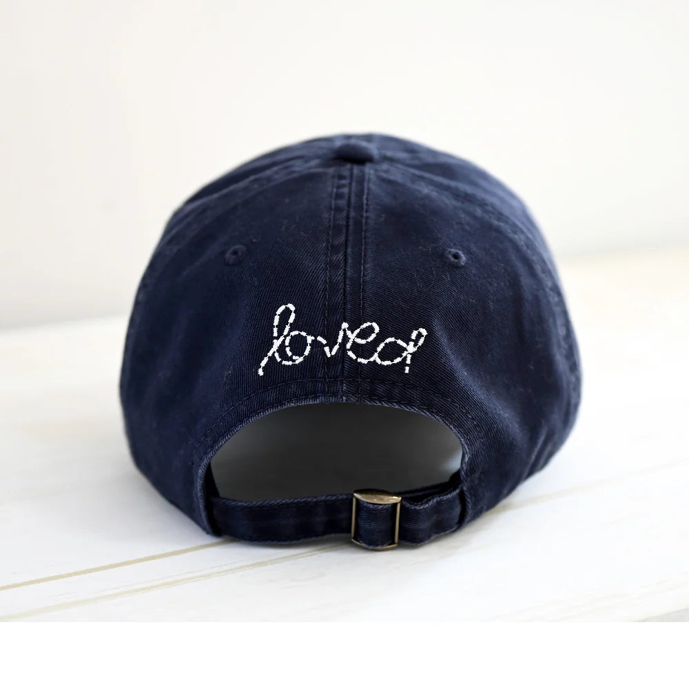 The Kerri Rosenthal Baseball Hat Heart Patch features &quot;loved&quot; and a white embroidered heart above the adjustable strap—a navy cap and chic essential, shown here on a light surface.