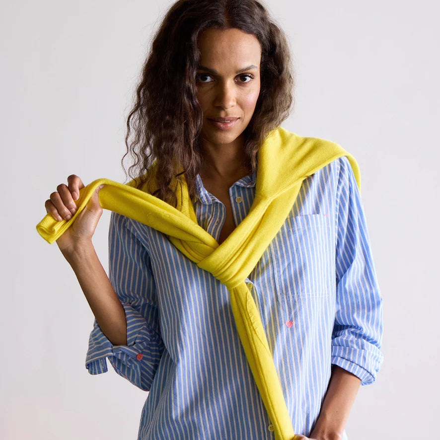 A person with long curly hair, wearing the Kerri Rosenthal Mia Shirt in stripe blue/white, has a yellow sweater draped over their shoulders while smiling slightly at the camera. The background is plain and light-colored.