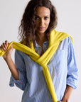 A person with long curly hair, wearing the Kerri Rosenthal Mia Shirt in stripe blue/white, has a yellow sweater draped over their shoulders while smiling slightly at the camera. The background is plain and light-colored.