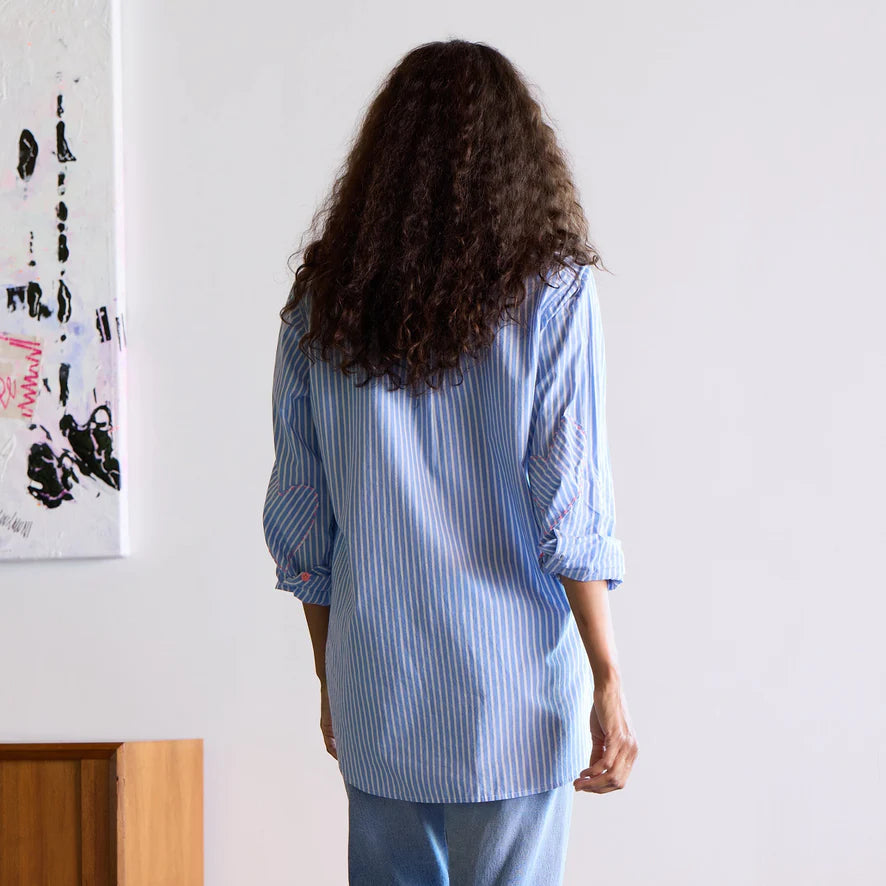 A person with curly hair is facing away, wearing the Mia Shirt Stripe Blue/White by Kerri Rosenthal paired with jeans. They are standing in a room adorned with white walls, a wooden cabinet, and partially visible abstract art on the wall.