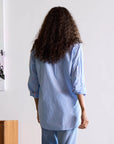 A person with curly hair is facing away, wearing the Mia Shirt Stripe Blue/White by Kerri Rosenthal paired with jeans. They are standing in a room adorned with white walls, a wooden cabinet, and partially visible abstract art on the wall.