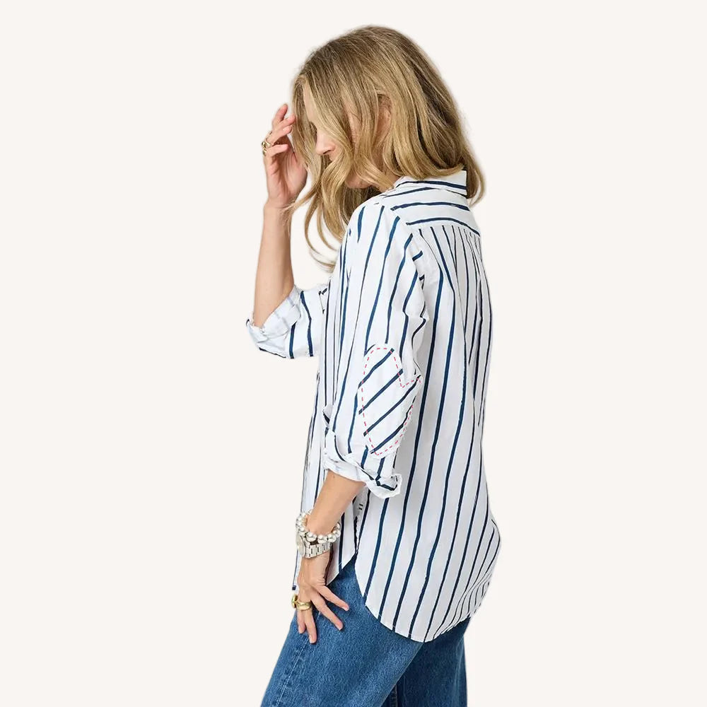 A woman stands sideways, touching her wavy blonde hair while wearing the Kerri Rosenthal Mia Mr Sharpie Stripe Shirt—a women’s cotton poplin shirt with vertical blue stripes and rolled-up sleeves—paired with blue jeans against a plain white background.