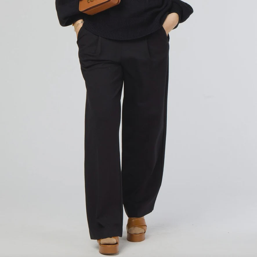 A person wearing a black sweater and Kerri Rosenthal's Wide Leg Trouser Pant in black stands against a plain background. The high-waisted pants exhibit a stylish flair, complemented by brown platform shoes. With hands in pockets, they casually carry a brown bag over their shoulder.