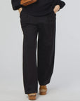 A person wearing a black sweater and Kerri Rosenthal's Wide Leg Trouser Pant in black stands against a plain background. The high-waisted pants exhibit a stylish flair, complemented by brown platform shoes. With hands in pockets, they casually carry a brown bag over their shoulder.