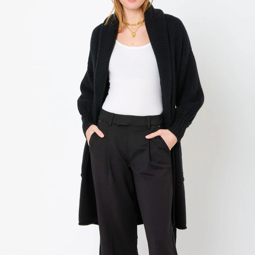 A person stands against a white background wearing a white top, black long cardigan, and Kerri Rosenthal's Wide Leg Trouser Pant in black. They have their hands in their pockets and are accessorized with gold necklaces.