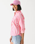 A woman in the Kerri Rosenthal Wide Stripe Pia pink and white cotton shirt, light blue denim shorts, and a pink baseball cap stands smiling in profile against a white background.