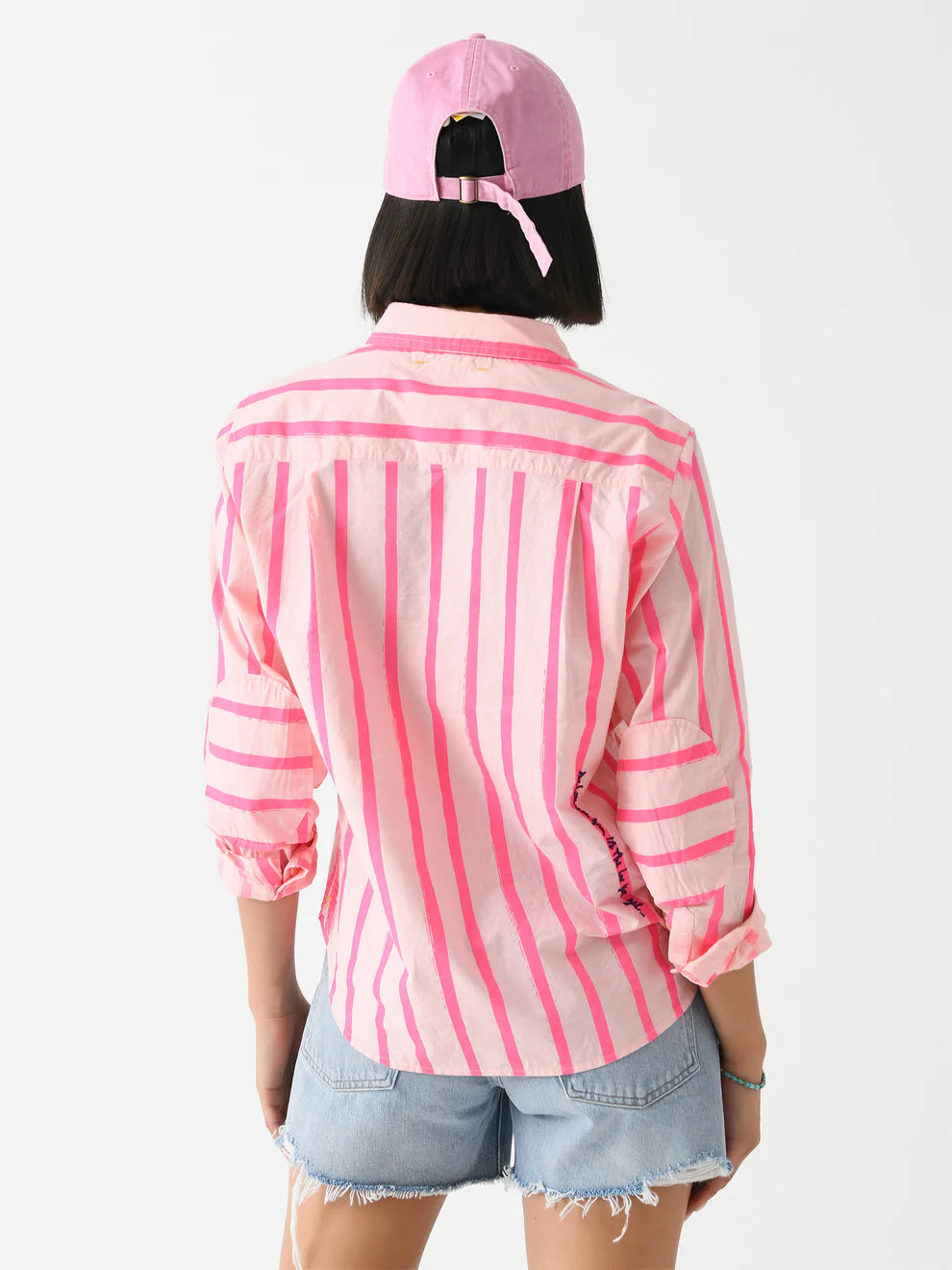 A person with a bob haircut faces away, wearing Kerri Rosenthal's Wide Stripe Pia relaxed-fit pink and white cotton shirt, sleeves rolled up. They pair it with light denim shorts and a pink baseball cap against a white background.