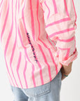 A person wears the Kerri Rosenthal Wide Stripe Pia shirt—pink and white stripes with handwritten text on the side—styled with rolled-up sleeves, light blue jeans, and a hand on hip for an effortless, laid-back look.