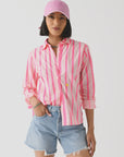 A woman with short dark hair and a relaxed, confident expression stands against a white background wearing the Kerri Rosenthal Wide Stripe Pia— a lightweight cotton pink and white striped shirt. She pairs it with light blue denim shorts and a pink baseball cap.