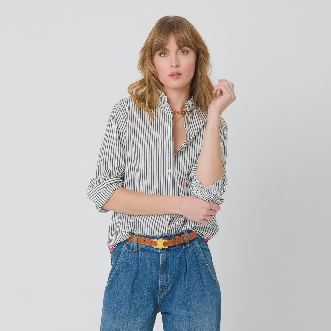 A woman with light brown hair is posing against a plain background. She is wearing a feminine Kerri Rosenthal Mia Ruffle Shirt with black vertical stripes, blue high-waisted jeans, and a brown belt with a gold buckle. Her left hand is touching her hair, and her right hand is at her side.