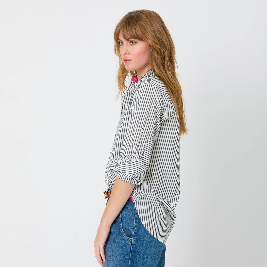 A person with long, wavy hair stands in profile against a plain background. They are wearing the Mia Ruffle Shirt by Kerri Rosenthal with the sleeves rolled up and blue jeans. Their expression appears neutral as they gaze to the side.