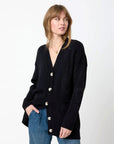 A person wearing a Kerri Rosenthal Poppy Zero Waste Cardigan in black, featuring imperfect hearts and pockets, paired with blue jeans, stands against a plain white background, gazing to the side.