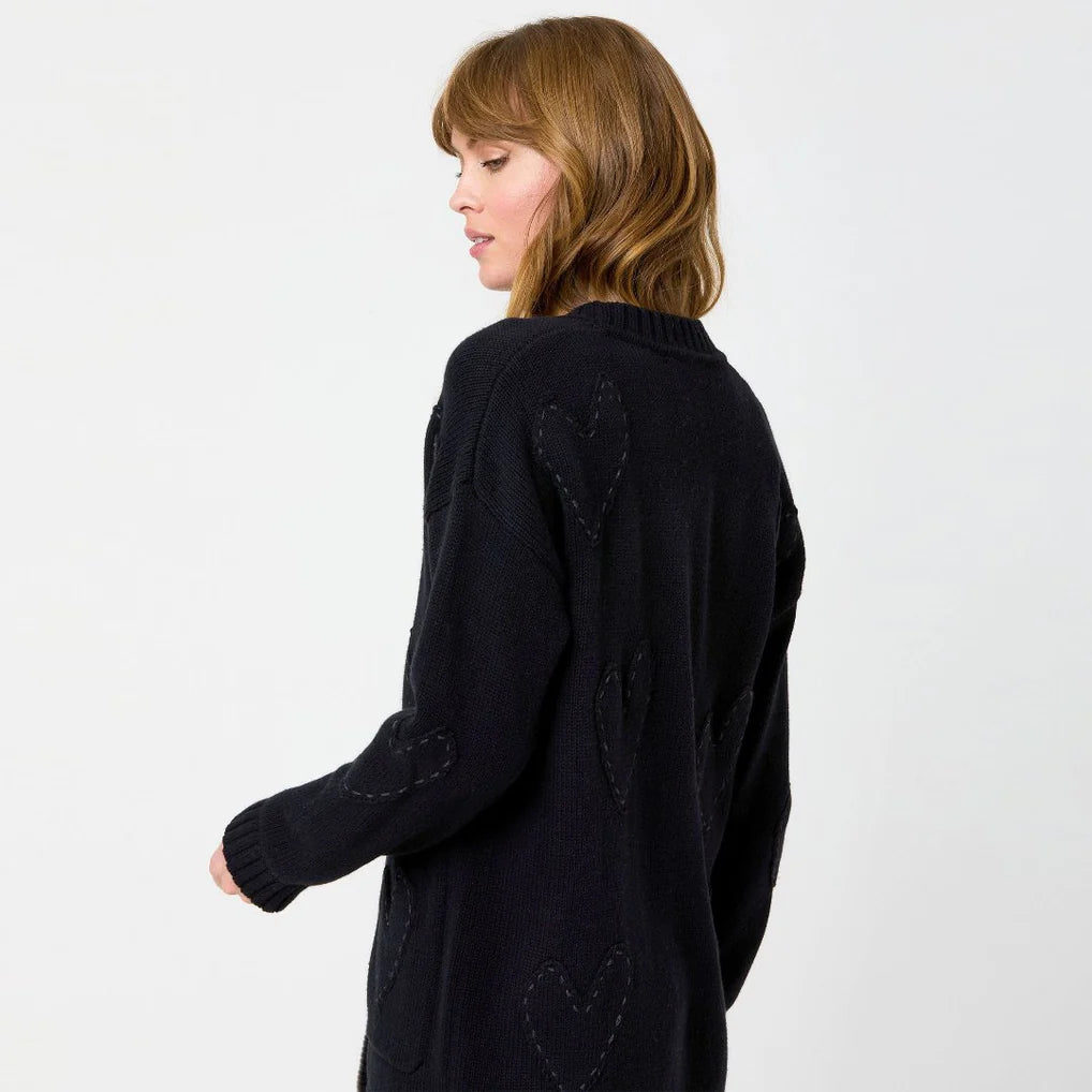 A woman with shoulder-length hair is wearing a black sweater decorated with imperfect hearts. She stands in a relaxed pose, embodying effortless style against a plain white background, reminiscent of the Kerri Rosenthal Poppy Zero Waste Cardigan.