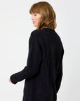 A woman with shoulder-length hair is wearing a black sweater decorated with imperfect hearts. She stands in a relaxed pose, embodying effortless style against a plain white background, reminiscent of the Kerri Rosenthal Poppy Zero Waste Cardigan.