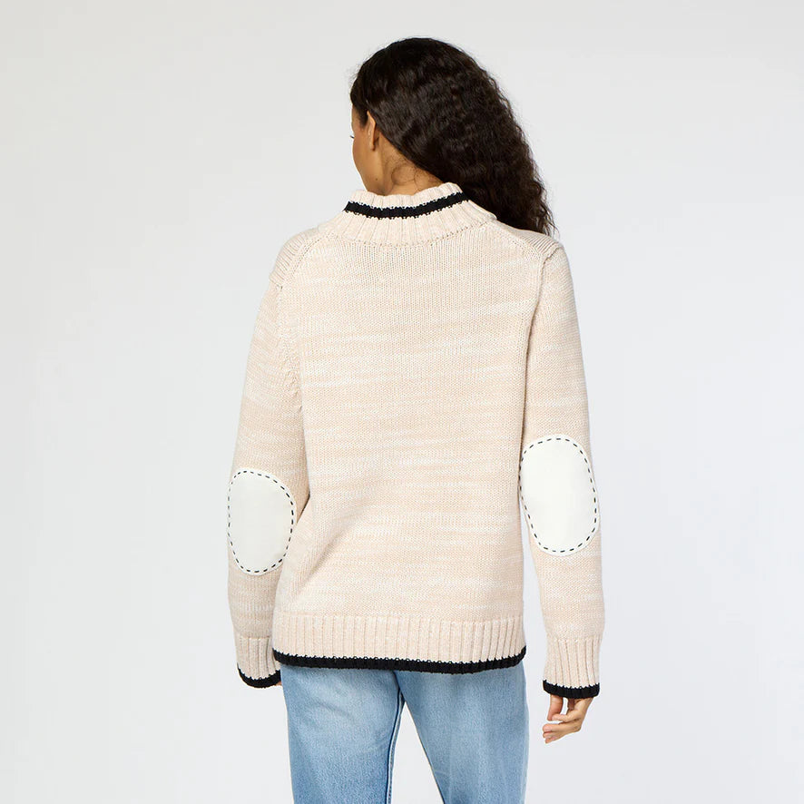 A rear view of an individual with long curly hair is shown wearing the Joy Love More Cardigan, a light beige knit sweater from Kerri Rosenthal featuring "Love More" embroidery and elbow patches. This is paired with light blue jeans as they stand against a plain background.