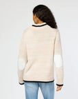 A rear view of an individual with long curly hair is shown wearing the Joy Love More Cardigan, a light beige knit sweater from Kerri Rosenthal featuring "Love More" embroidery and elbow patches. This is paired with light blue jeans as they stand against a plain background.