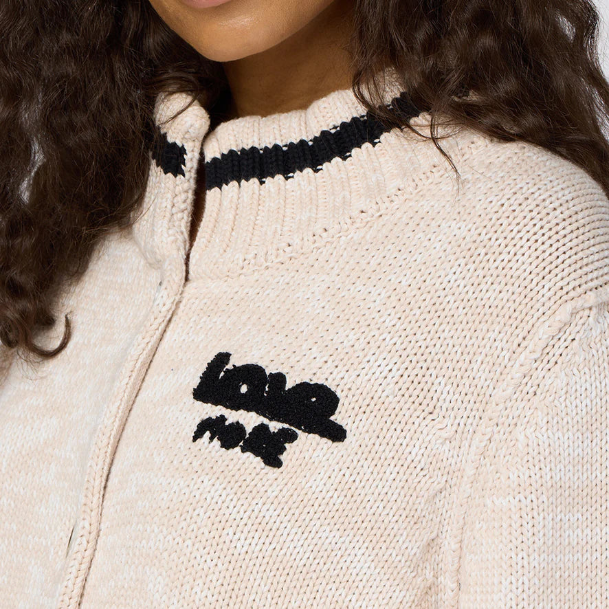 The individual wears a beige knit sweater adorned with a black and white striped collar, beautifully embroidered with "love more" on the chest. This cozy piece, reminiscent of the Joy Love More Cardigan by Kerri Rosenthal, pairs perfectly with their long, curly hair for an effortlessly chic look.