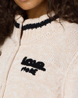 The individual wears a beige knit sweater adorned with a black and white striped collar, beautifully embroidered with "love more" on the chest. This cozy piece, reminiscent of the Joy Love More Cardigan by Kerri Rosenthal, pairs perfectly with their long, curly hair for an effortlessly chic look.