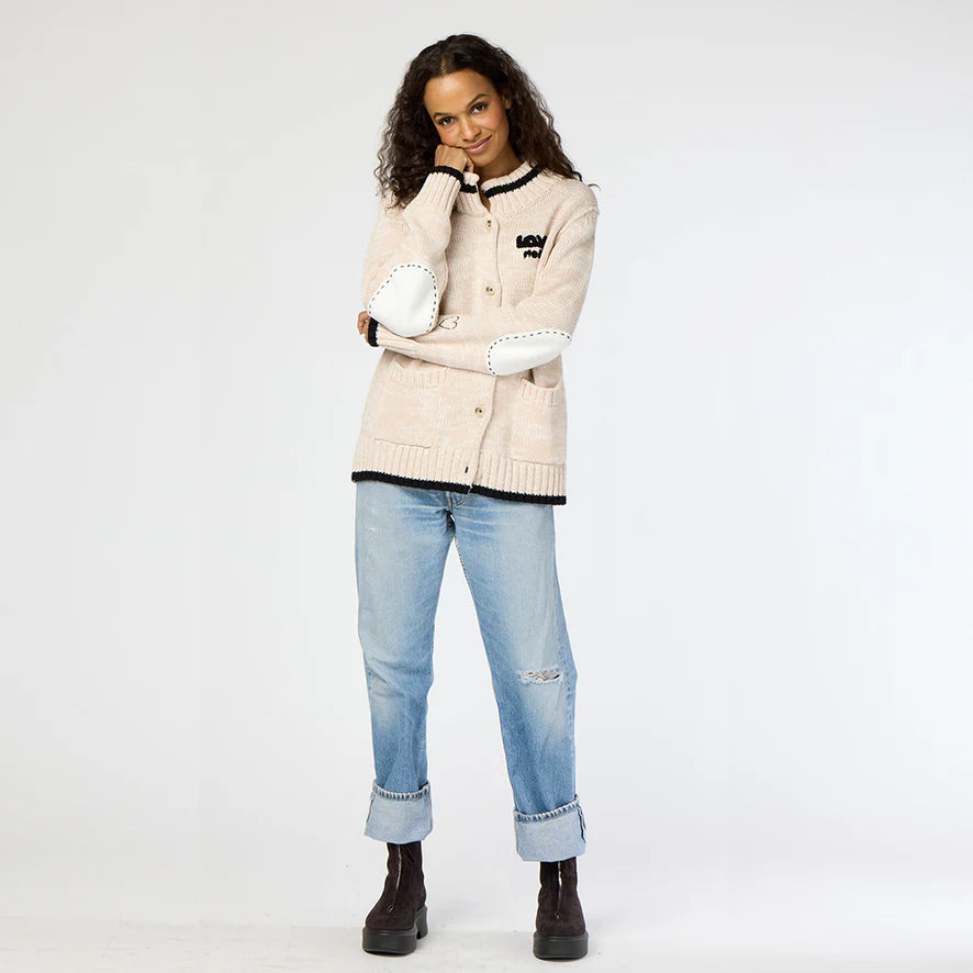 A person stands posing with one hand touching their chin, showcasing the Joy Love More Cardigan from Kerri Rosenthal. This piece is crafted from 100% cotton and features exquisite "Love More" embroidery. They pair it stylishly with blue rolled-up jeans and dark boots against a plain white background.