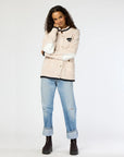 A person stands posing with one hand touching their chin, showcasing the Joy Love More Cardigan from Kerri Rosenthal. This piece is crafted from 100% cotton and features exquisite "Love More" embroidery. They pair it stylishly with blue rolled-up jeans and dark boots against a plain white background.