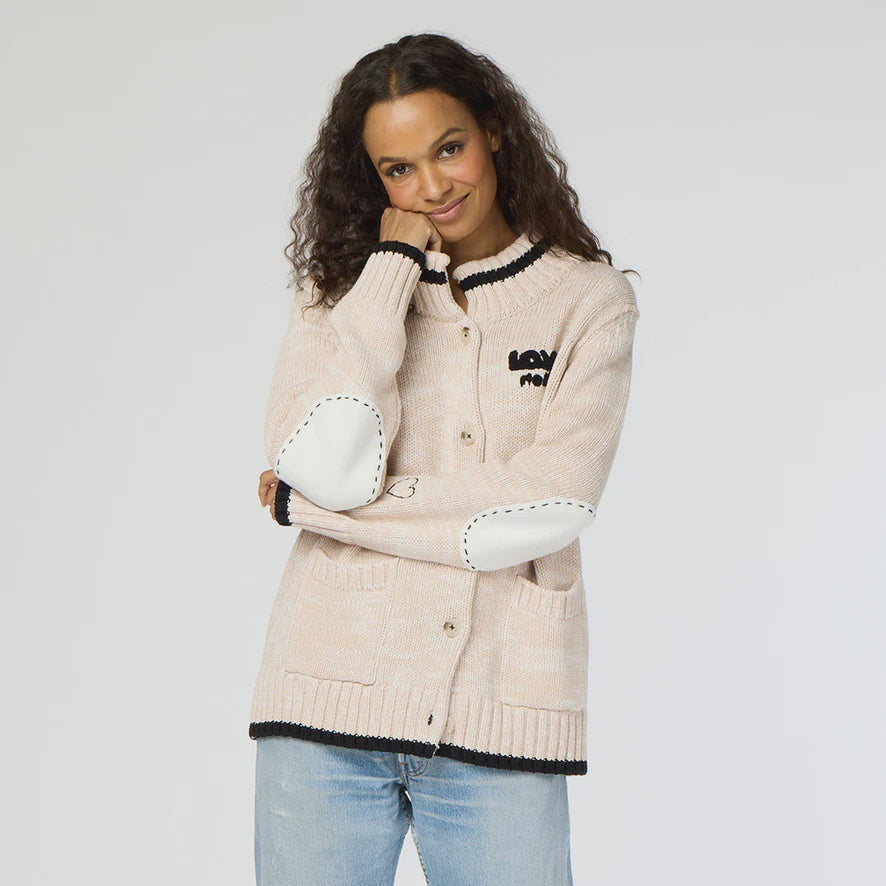 A person with long curly hair wears the Kerri Rosenthal Joy Love More Cardigan—a light beige button-up with black trim and white elbow patches, crafted from 100% cotton. Paired with blue jeans, they are smiling and standing against a plain background.