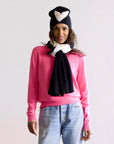 A person wearing a bright pink sweater, light blue jeans, and a black and white scarf stands against a plain background. Their black beanie showcases the Heart Beanie design by Kerri Rosenthal, crafted with soft cotton/cashmere. They have long, dark hair and a relaxed posture, smiling gently. Enjoy free shipping!