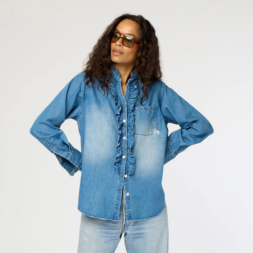 A person wearing sunglasses and the Kerri Rosenthal Molly Denim Shirt with ruffle trim, paired with lightweight denim jeans, confidently stands with hands on hips against a plain light background.